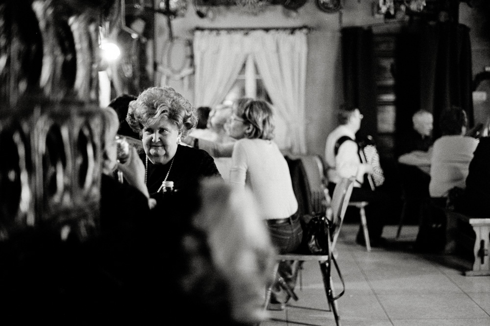 Schwarz-Weiß-Foto einer älteren Frau mit voluminöser Frisur sitzt an einem Tisch in einem belebten Wirtshaus.