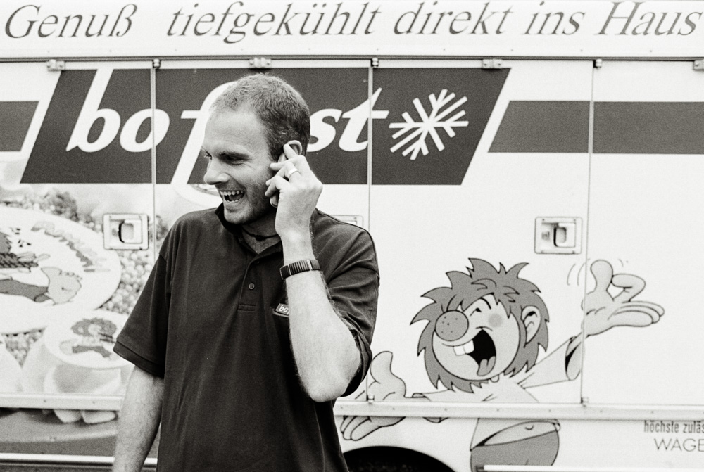 Schwarz-Weiß-Foto eines jungen Mannes lacht am Telefon vor einem bofrost-Lieferwagen mit aufgemalter Zeichentrickfigur.