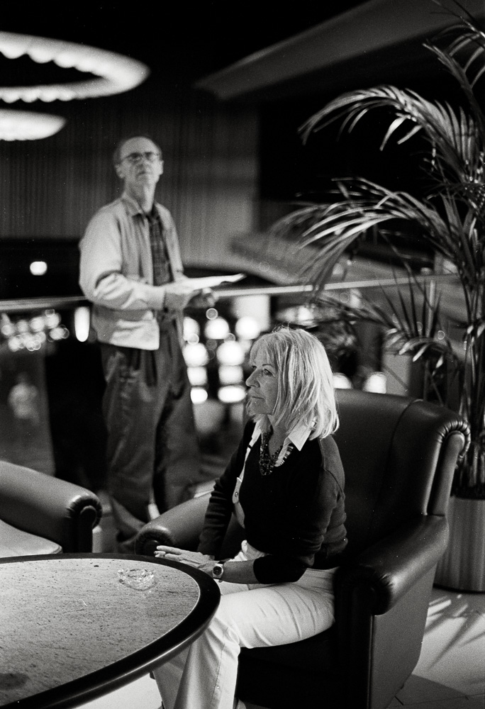 Schwarz-Weiß-Foto von zwei Personen in einer Lounge: eine Frau sitzt im Ledersessel, ein älterer Mann blickt von einer Terrasse.
