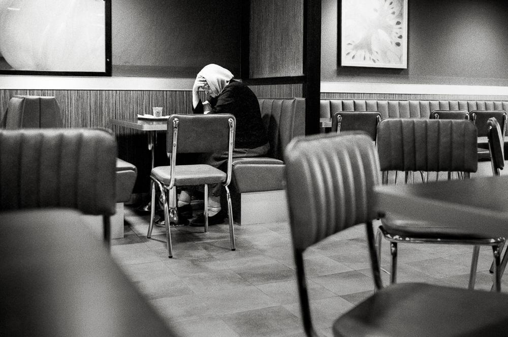 Schwarz-Weiß-Foto einer erschöpften Person sitzt zusammengesunken mit gesenktem Kopf an einem Tisch in einem Imbiss.
