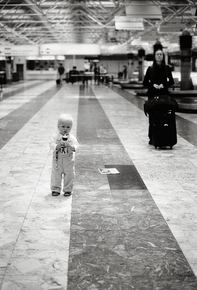 Schwarz-Weiß-Foto eines Kleinkinds mit Flasche an der Gepäckausgabe am Flughafen, dahinter eine nach oben blickende Frau.
