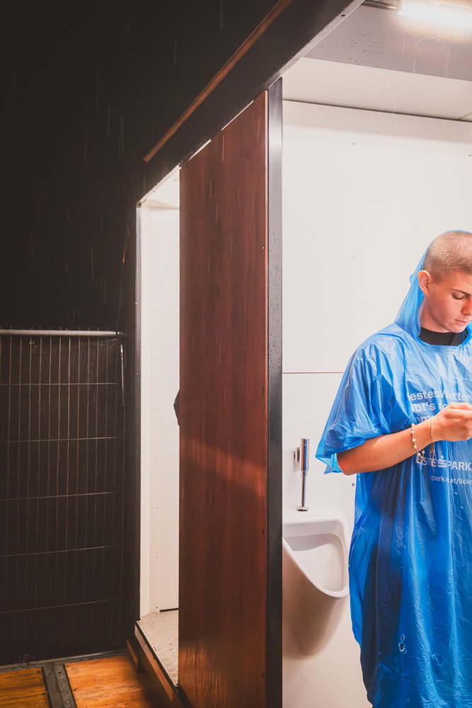 Ein Jugendlicher im blauen Regenponcho steht vor einer Mobiltoilette und tippt auf sein Handy.