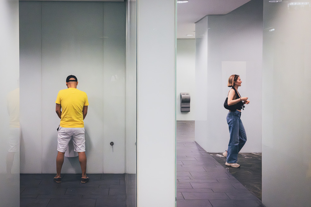Ein Mann steht an einer Toilettenwand auf der linken Seite, eine Frau geht im rechten Gang.