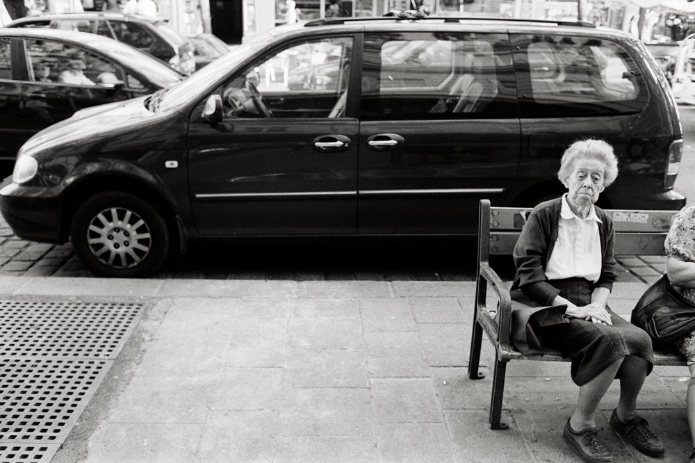 A black and white photo of an older woman with short hair sitting alone on a wooden sidewalk bench. She has a contemplative expression. A dark minivan is parked directly behind her.