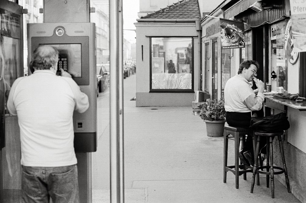 A black and white street scene. On the left, a man is standing and using an ATM. On the right, another man sits on a barstool at a small curbside food stand, eating a hot dog.