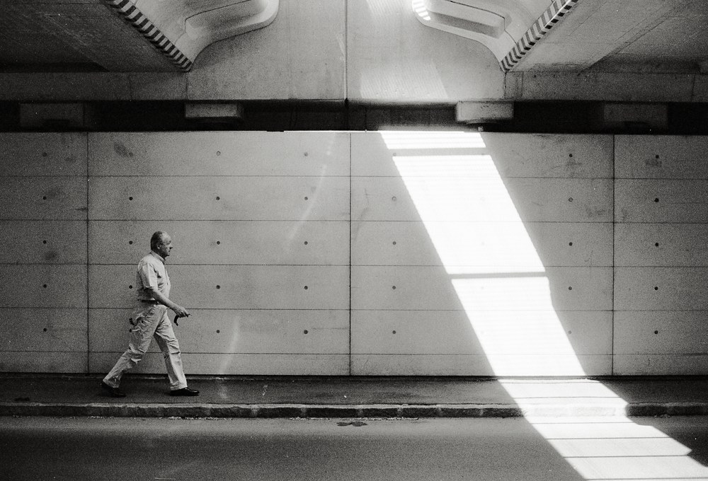 A black and white photo of a man walking along a sidewalk beneath a large concrete overpass or bridge. A sharp, bright diagonal beam of sunlight cuts across the textured concrete wall in front of him.