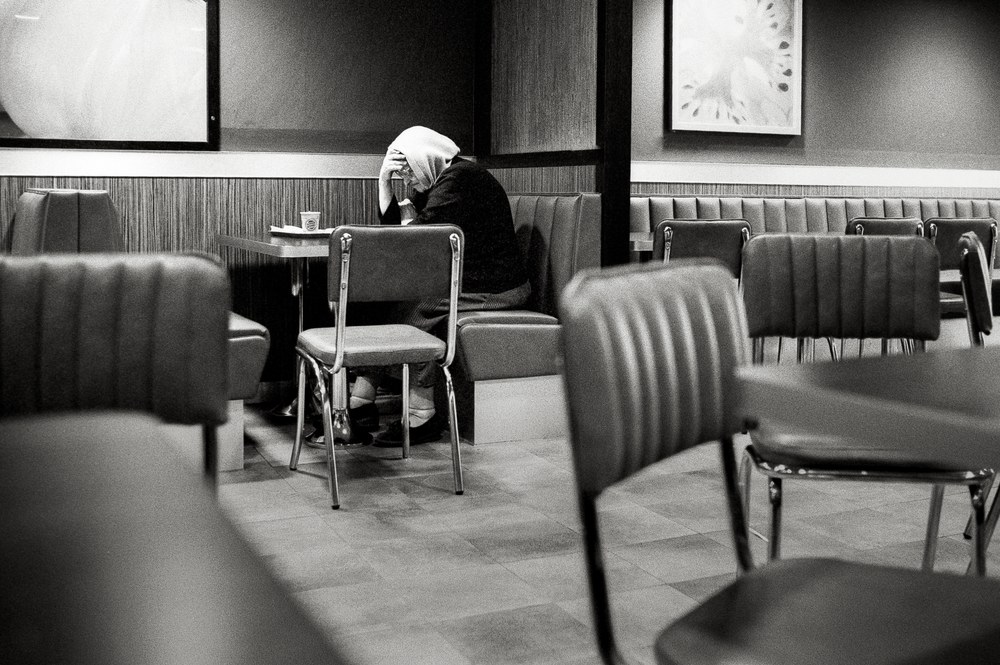 Schwarz-Weiß-Foto einer erschöpften Person sitzt zusammengesunken mit gesenktem Kopf an einem Tisch in einem Imbiss.