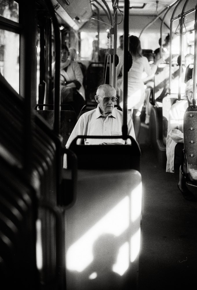 Schwarz-Weiß-Foto eines älteren Mannes mit Brille sitzt ruhig in einem Bus, während ein scharfer Lichtstrahl den Sitz vor ihm erhellt.