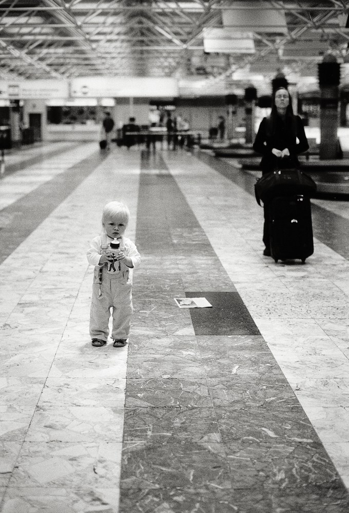 Schwarz-Weiß-Foto eines Kleinkinds mit Flasche an der Gepäckausgabe am Flughafen, dahinter eine nach oben blickende Frau.