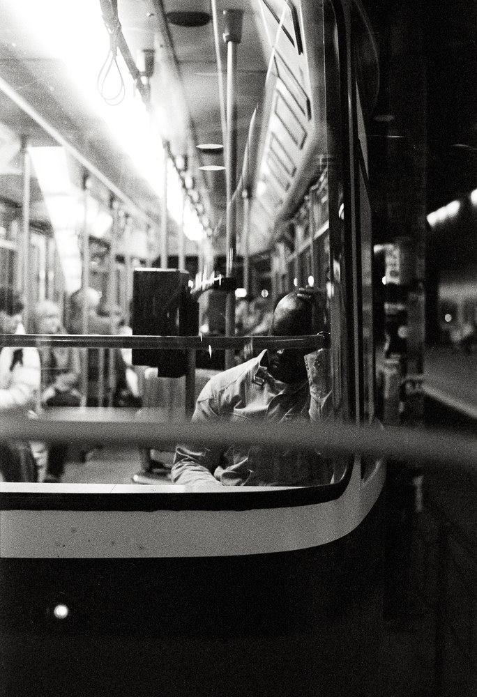 A black and white photo taken from outside a brightly lit tram at night, looking in through the window. Inside, a passenger is seated with their head resting heavily on their hand, appearing tired or lost in thought.