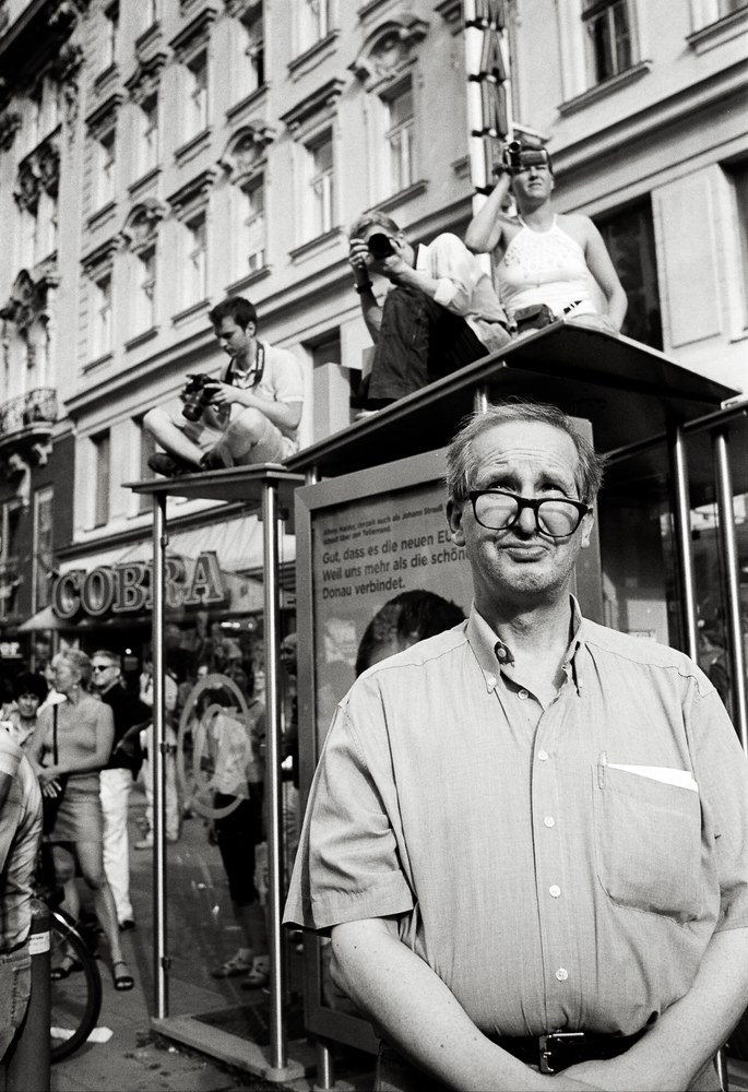 Schwarz-Weiß-Porträt eines älteren Mannes mit dicker Brille im Vordergrund. Dahinter sitzen zwei Personen auf einem gläsernen Bushaltestellendach.