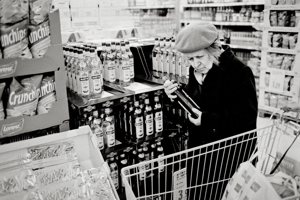 A black and white photo of an older woman wearing a beret and a dark coat in a grocery store aisle. She is holding a bottle of liquor like you would a child, ready to place it into the shopping cart parked in front of her.