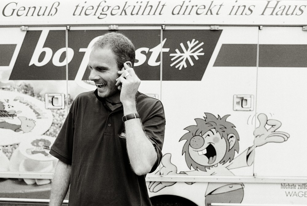 A black and white photo of a young man laughing while talking on a cell phone. He is standing in front of a "bofrost" delivery truck, which features a large, expressive cartoon character painted on the side.