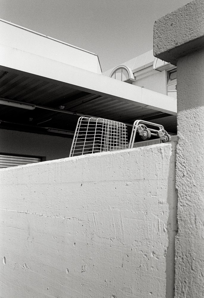 A black and white photo looking up at a thick, textured concrete wall. An empty wire shopping basket rests on top of the wall, with the flat awning and roof of a modern commercial building visible behind it.