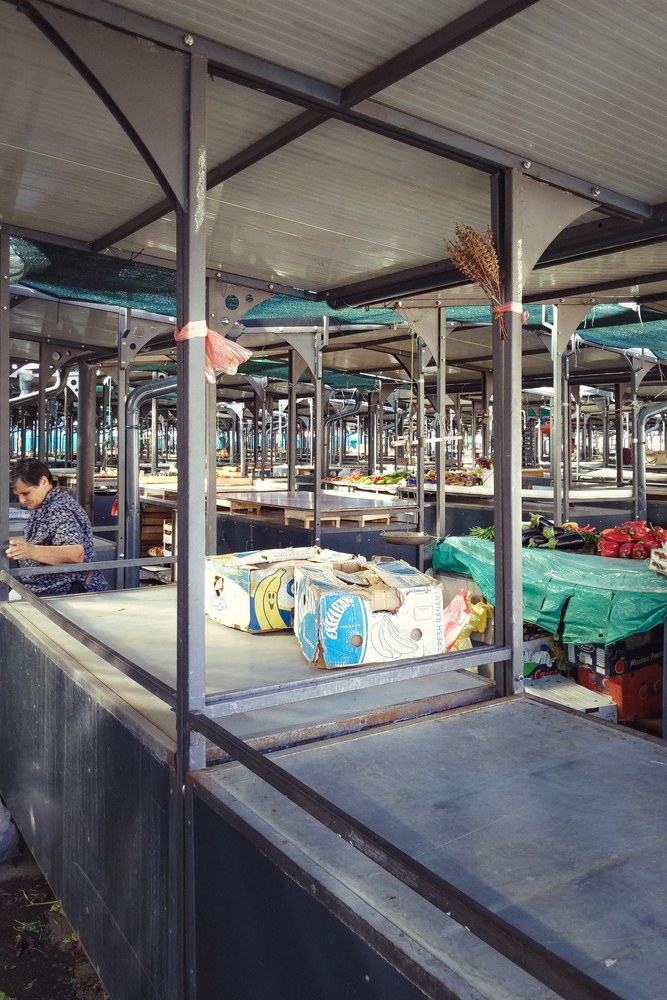 A woman sits behind a metal stall in a wholesale indoor market, looking down at her hands, next to a cardboard box of bananas and covered produce.