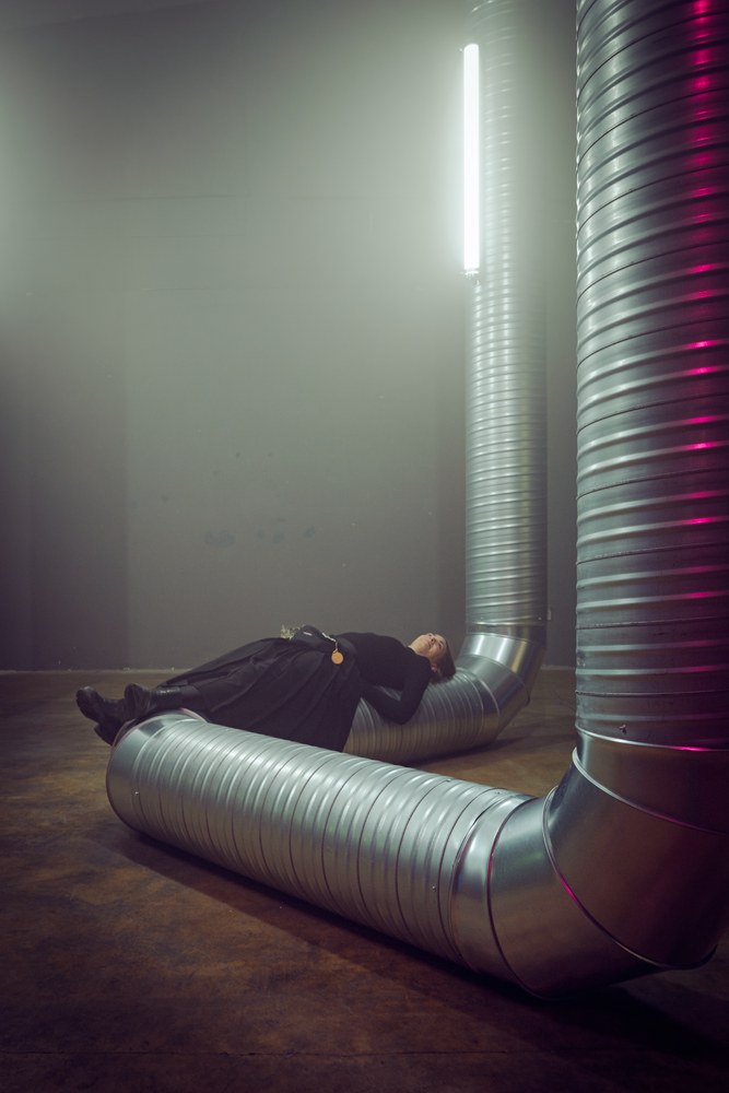 An industrial-looking room with a concrete floor and dark grey walls. A large, ribbed silver ventilation duct curves across the floor. A person dressed entirely in black is lying horizontally, resting their back directly across the metal duct beneath a bright fluorescent light.
