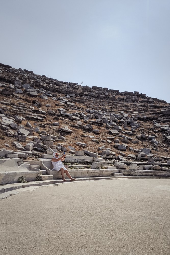 Eine Frau in weißem kleid und breitkrempigem weißem Hut sitzt auf den steinstufen eines antiken Amphitheaters und hält ein Handy hoch, als würde sie einen Selfie machen.