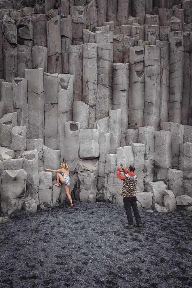 Ein Farbfoto eines schwarzen Sandstrands mit hohen Basaltsäulen. Eine Frau in einem hellblau-weißen Badeanzug posiert barfuß am Felsen, während ein Mann in oranger Jacke und Tarnweste sie mit einem Smartphone fotografiert.