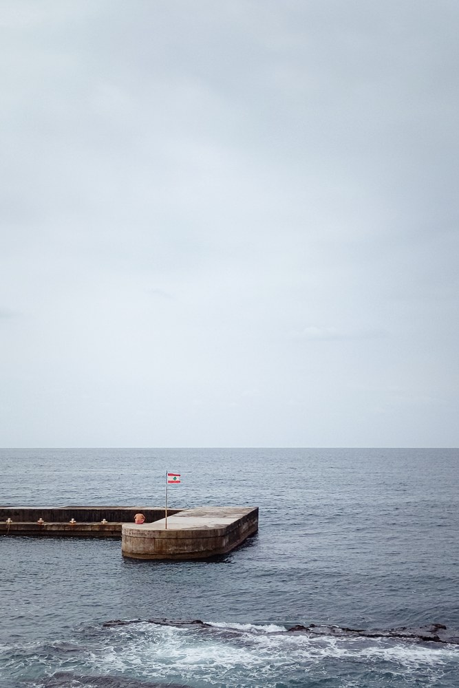 Eine Person sitzt am Pier vor dem ruhigen Meer neben einer wehenden libanesischen Flagge.