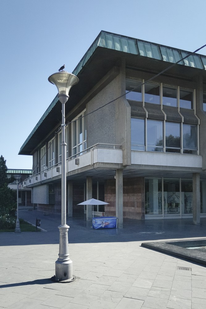 A slightly faded color photo of a large concrete building with a green copper roof edge. In the foreground, a pigeon sits perched on top of a tall, classic streetlamp. On the paved plaza beneath the building, there is a small blue ice cream stand with a white umbrella.